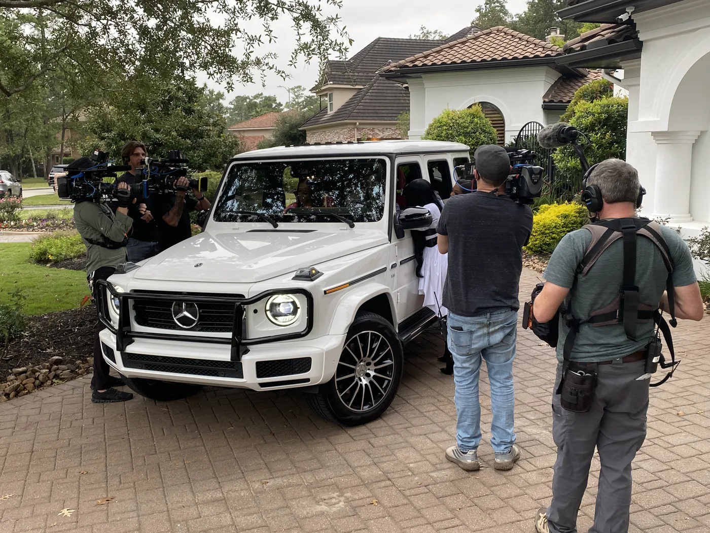 TV crew filming a white SUV in the driveway of a large home